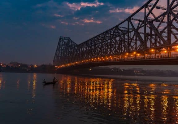 A breathtaking night view of the iconic Howrah Bridge in Kolkata, India. The bridge is illuminated with warm lights, reflecting on the calm waters of the Hooghly River. A small boat with a lone rower adds to the picturesque scene.