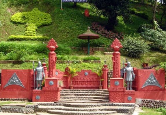 A view of the Agnigarh Fort in Assam, India. The image showcases the fort's unique architecture with red stonework, intricate carvings, and landscaped gardens.