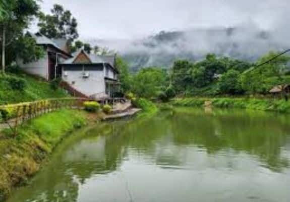 A scenic view of a resort with cottages nestled amidst lush greenery and a calm lake in the foothills of the Himalayas.