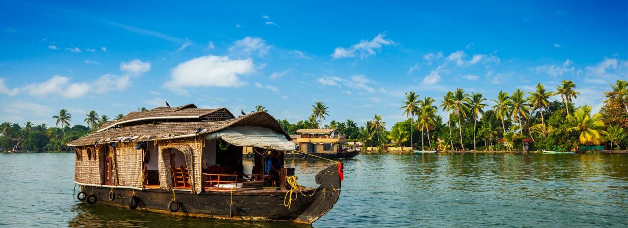 A traditional houseboat, known as a Kettuvallam, is moored on the tranquil backwaters of Kerala, India. Lush green palm trees line the shore, and a clear blue sky completes the idyllic scene.