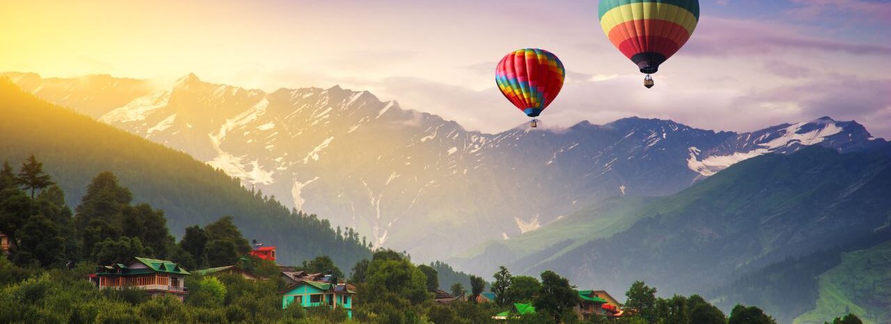 Two colorful hot air balloons soar over a picturesque valley in Manali, Himachal Pradesh, India. Snow-capped mountains form a stunning backdrop to this idyllic scene.